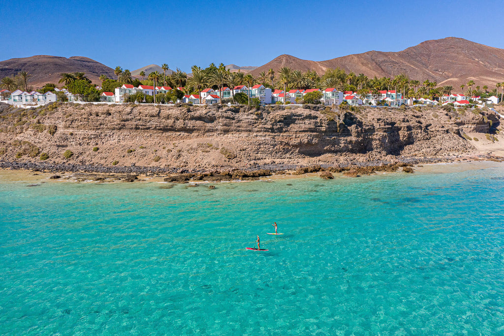 Two stand-up paddleboarders in clear turquoise water with coastal village and volcanic mountains in background, Fuerteventura.
