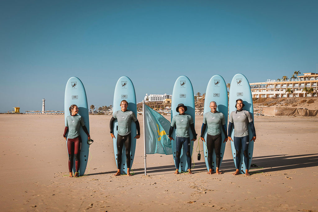 5 Instructors from Surfers Island standing with boards t the beach of Morro Jable in front of the Surfers Island Station with waving flag of Surfers Island