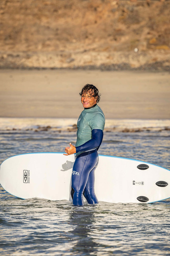 Surfinstructor Vincent standing in the water, holding a surfboard giving the shaka hand sign